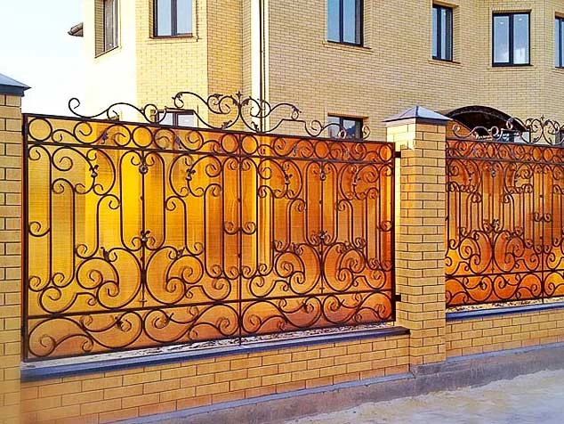 Забор с художественной ковкой из поликарбоната на кирпичных столбах для частного дома